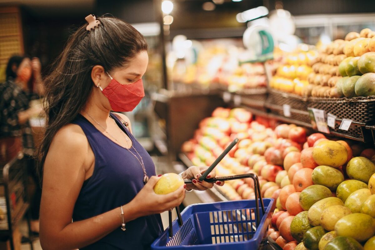 A woman wearing a mask selects fresh fruits in a supermarket produce aisle.